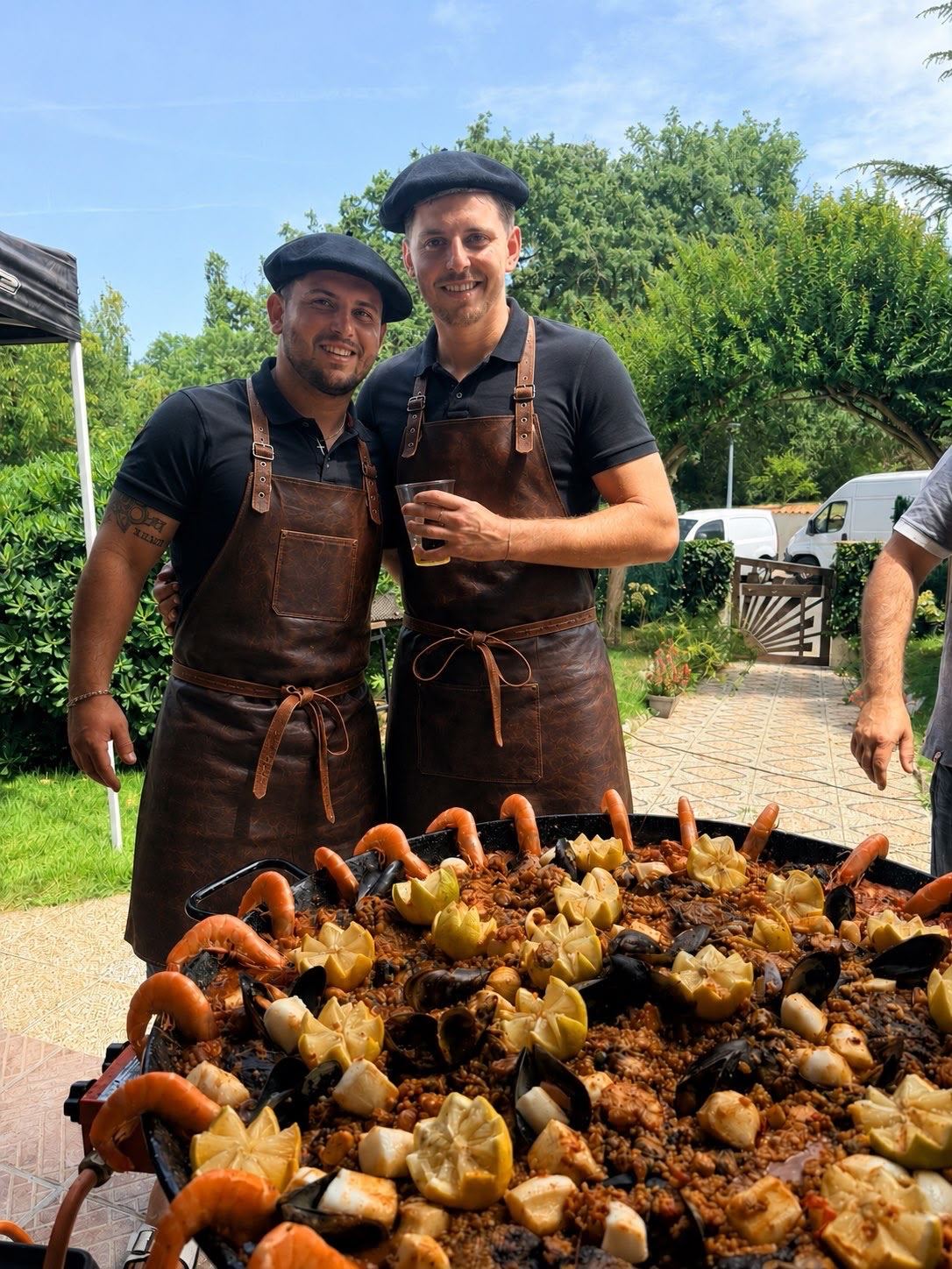 Les deux cuisiniers de La Paella des Copains, en tablier, souriants devant une grande paella